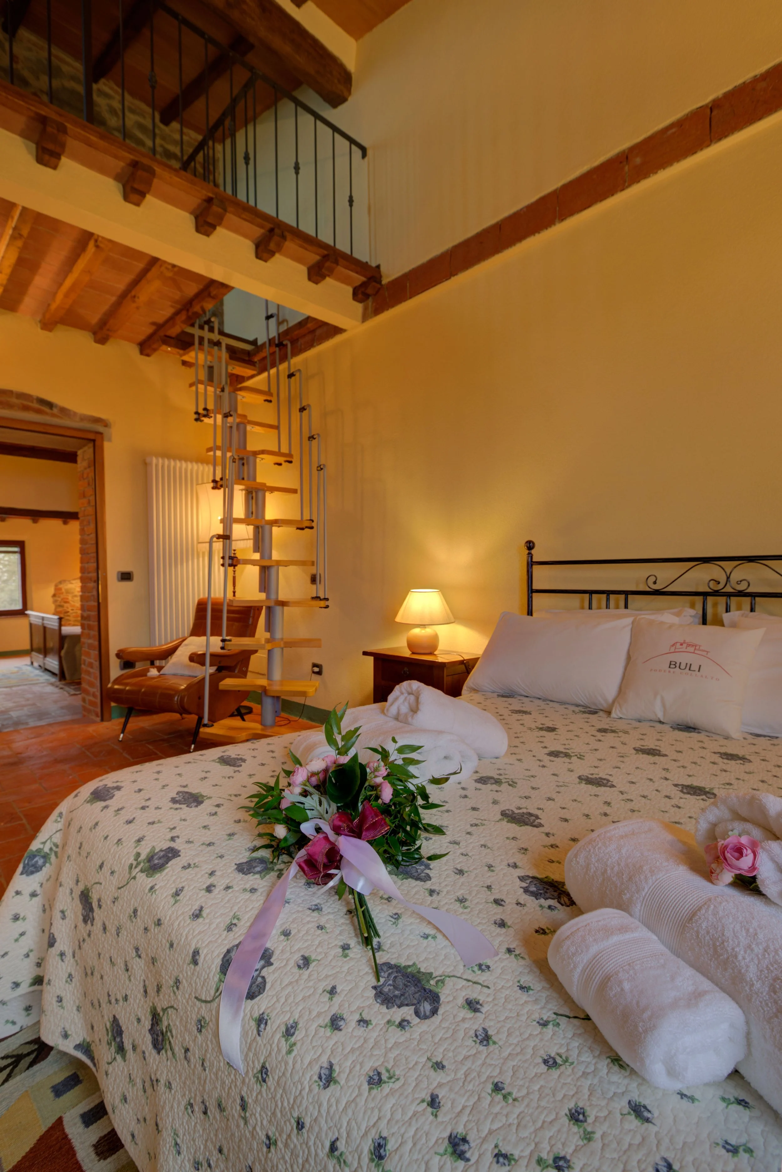 A cozy bedroom with a bed covered in a floral quilt, a bouquet of pink and white flowers on the bed, and two rolled white towels. A nightstand with a lamp is beside the bed. The room has warm yellow walls, exposed brick, and wooden ceiling beams. A s