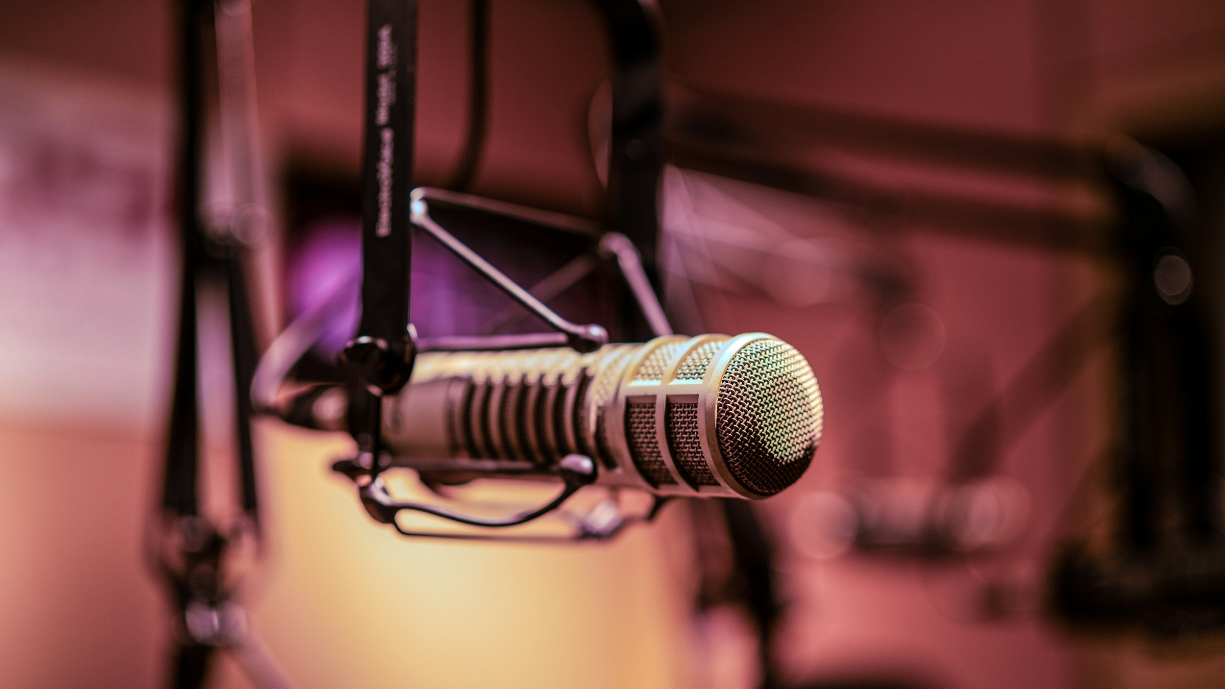 A professional microphone on a stand, with a blurred background of pink and purple lighting, likely in a recording studio.