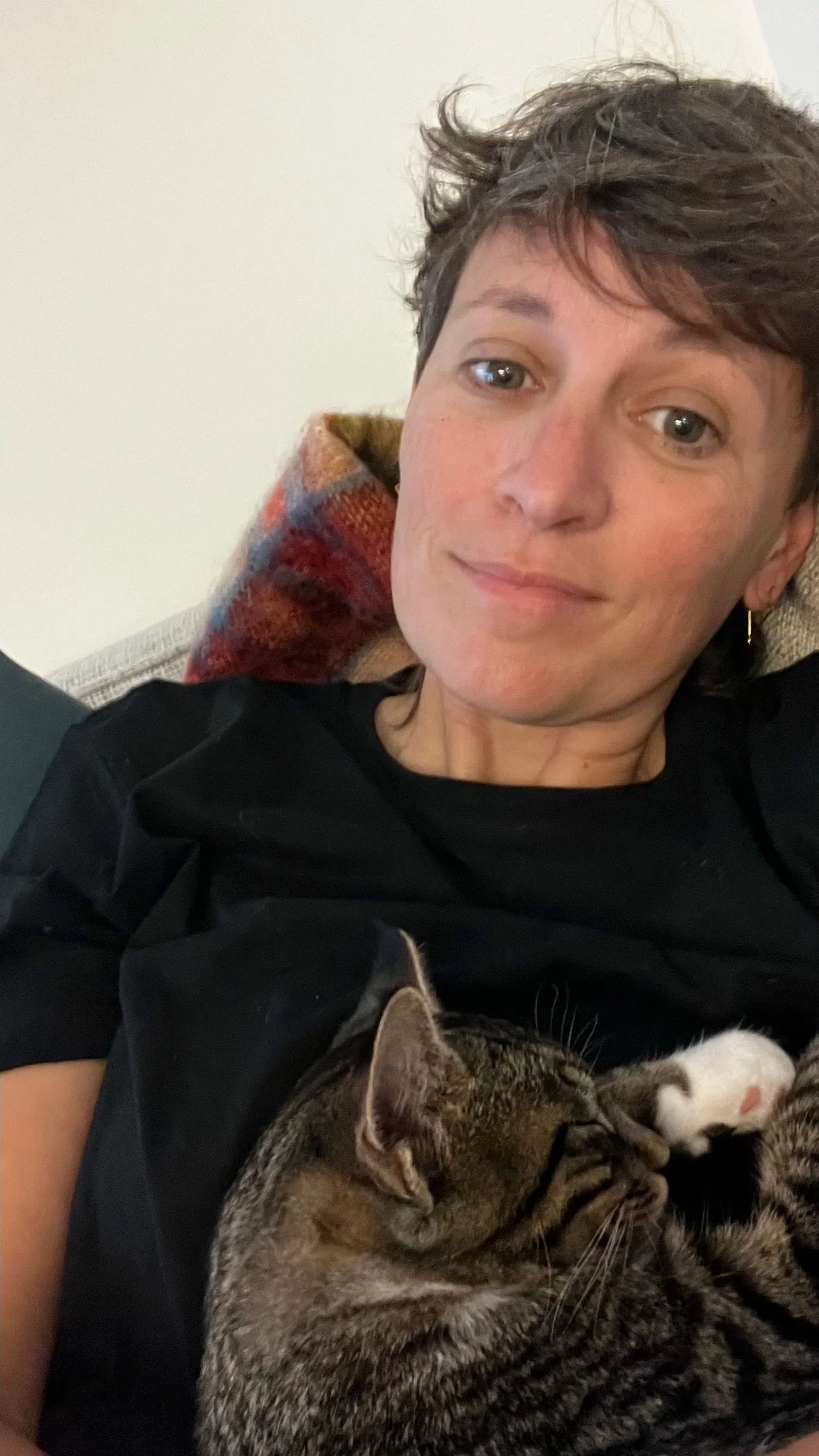 A person with short brown hair and light skin relaxing on a couch, with a tabby and a black-and-white cat cuddling on her chest.