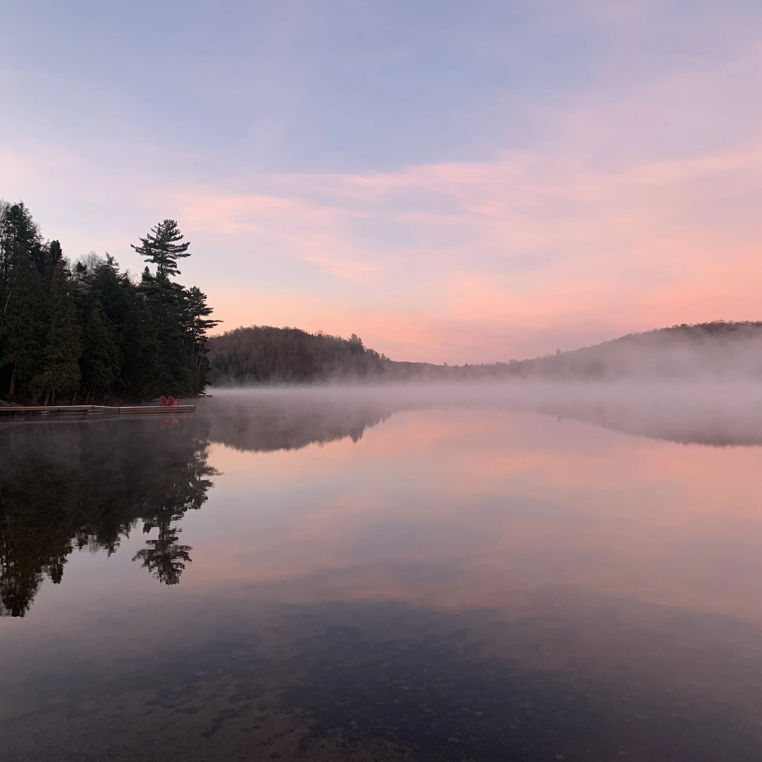 A serene lake at dawn reflecting a colorful sky with pink and purple clouds, surrounded by trees and hills in the distance.
