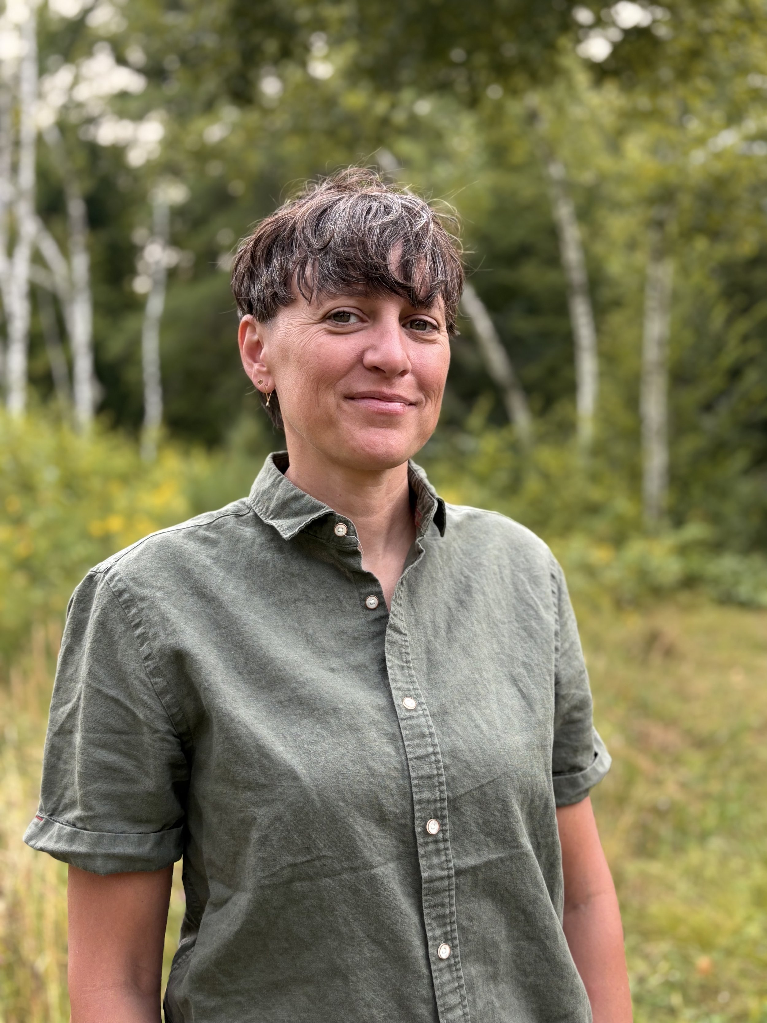 A person with short, wavy, slightly gray hair, wearing a button-up green shirt, standing outdoors in a forested area with birch trees in the background.