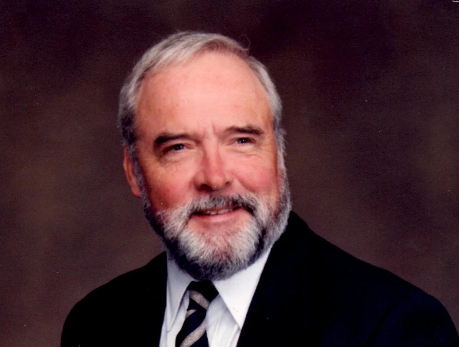Portrait of a smiling older man with gray hair and a beard, dressed in a black suit, white shirt, and striped tie, against a dark background.