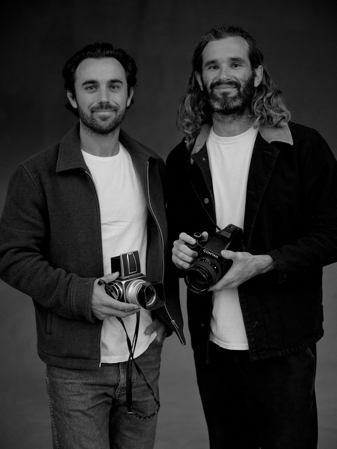 Two men standing side by side, each holding a vintage camera, smiling at the camera. The photo is in black and white.