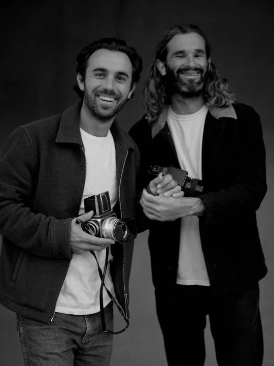 Two men smiling and holding cameras, standing side by side against a plain background.