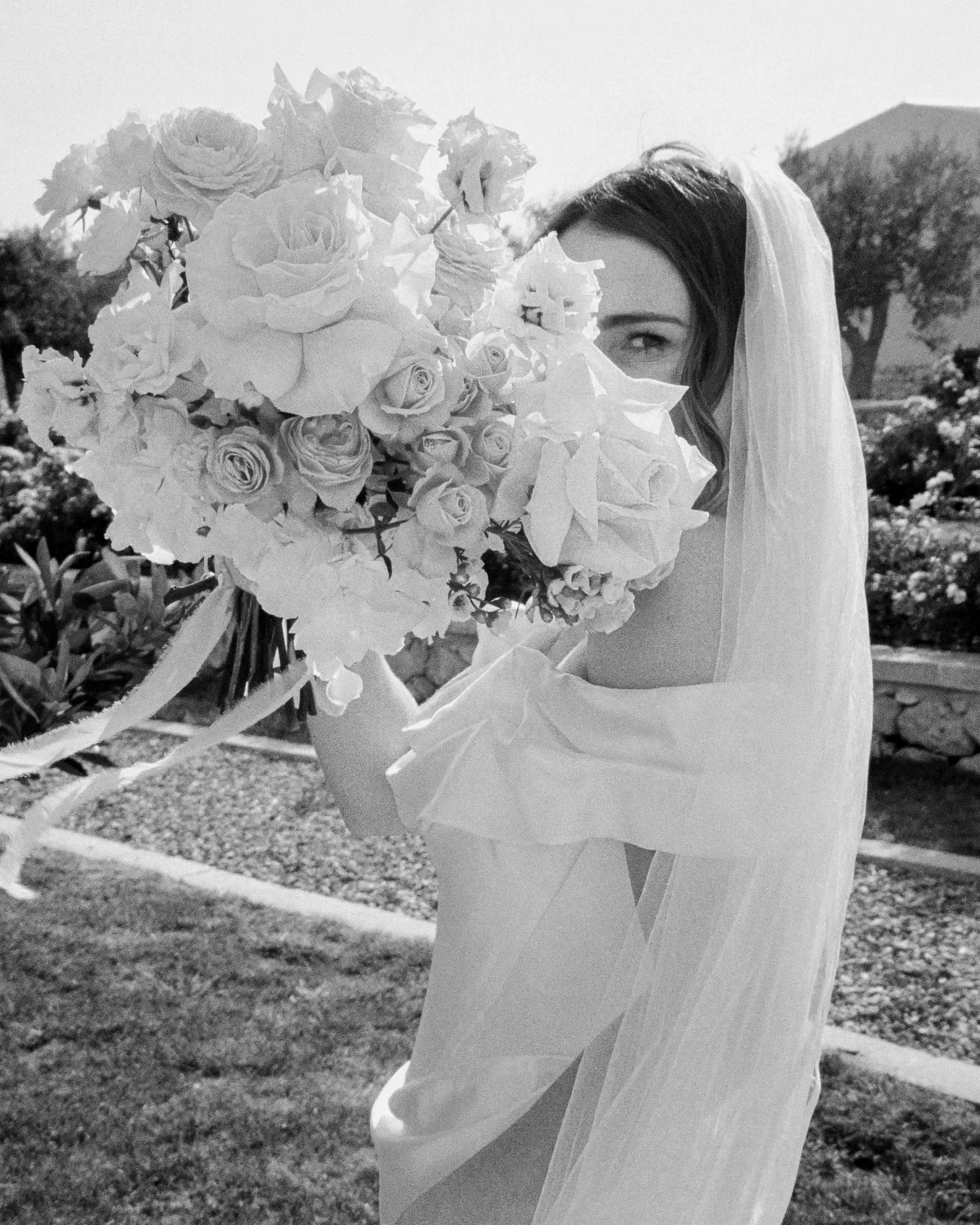 A bride in a wedding dress holds a large bouquet of roses and other flowers, partially hiding her face, outdoors in a garden or park setting.