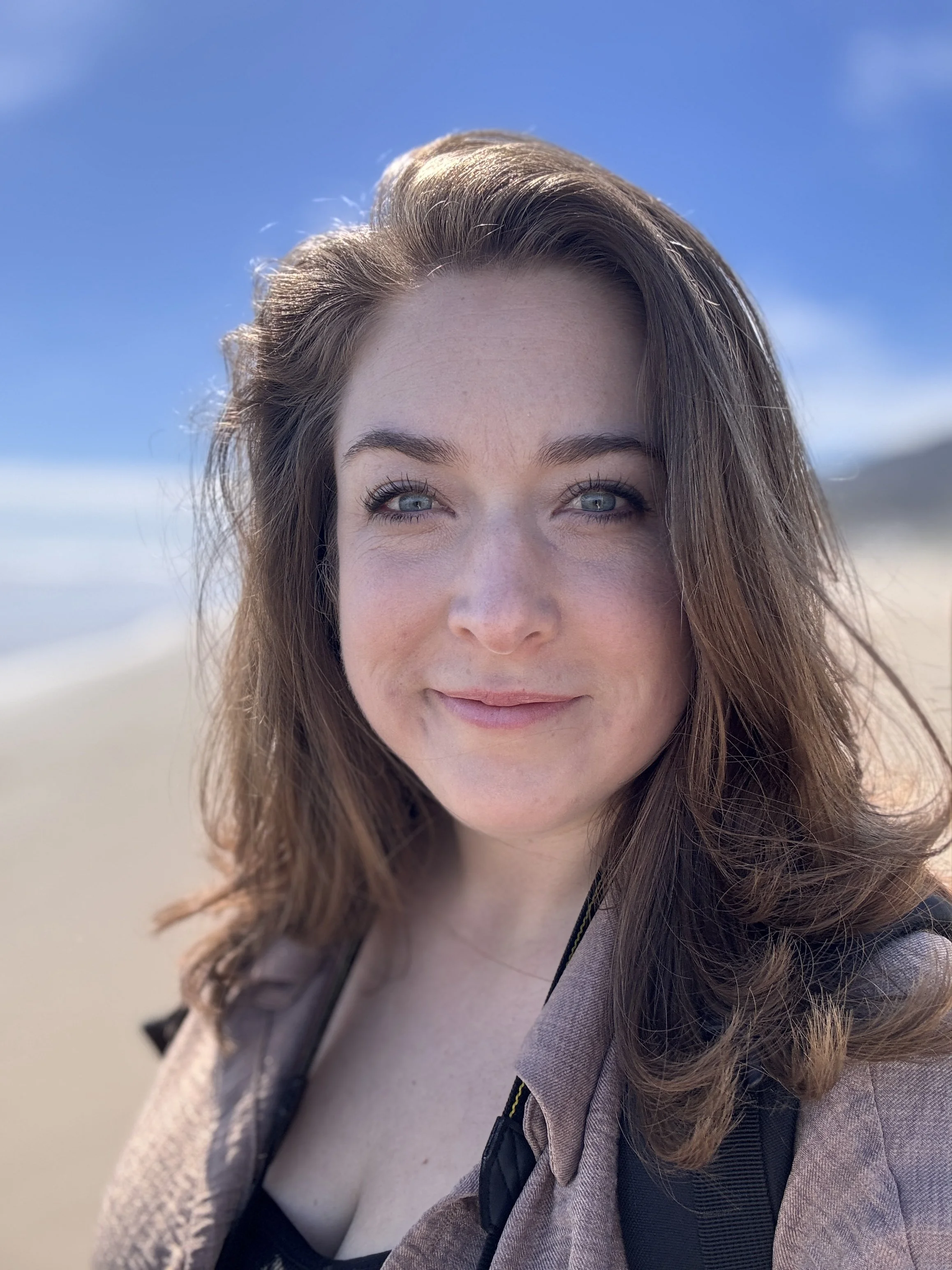 A woman with light brown hair and blue eyes smiling at the camera, outdoors with sandy ground and blue sky in the background.