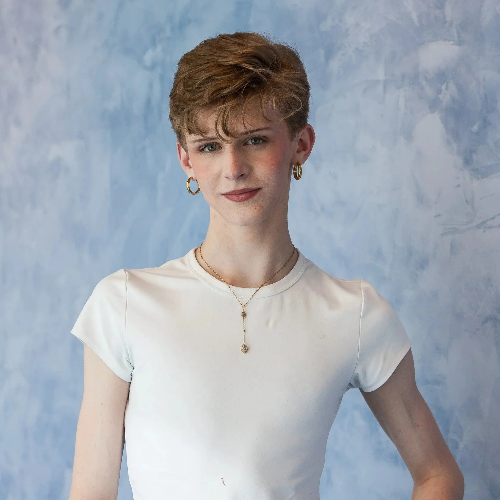 A woman with short, tousled hair, wearing gold hoop earrings, a delicate gold necklace, and a white short-sleeve top, standing against a blue textured background.
