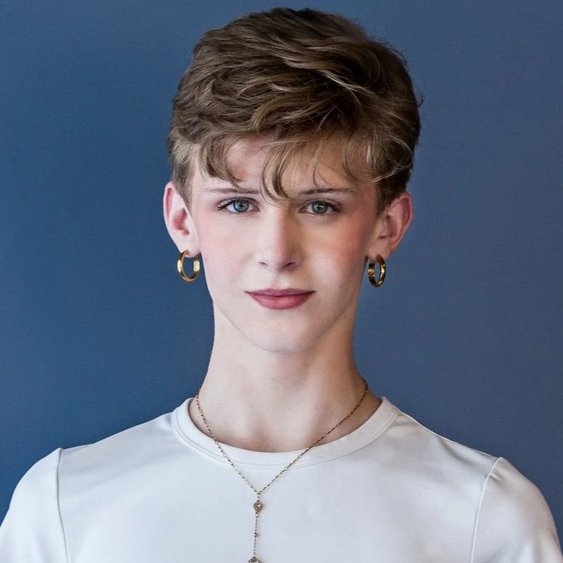 A portrait of a young person with short, wavy light brown hair and blue eyes, wearing a white top, gold hoop earrings, and layered necklaces, standing against a dark blue background.