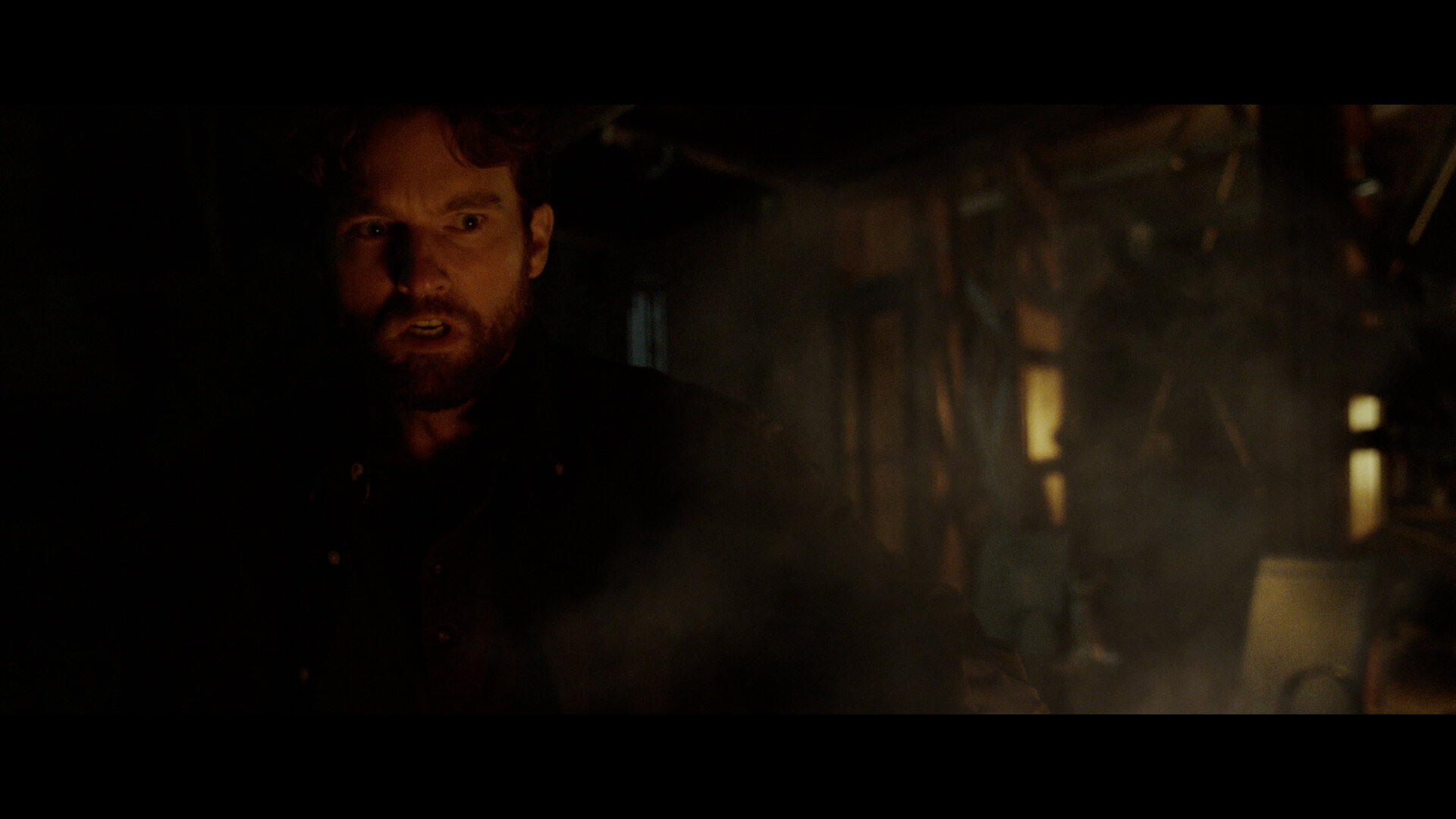 A man with a beard in a dark, smoky environment, illuminated by dim amber light, with a tense facial expression.
