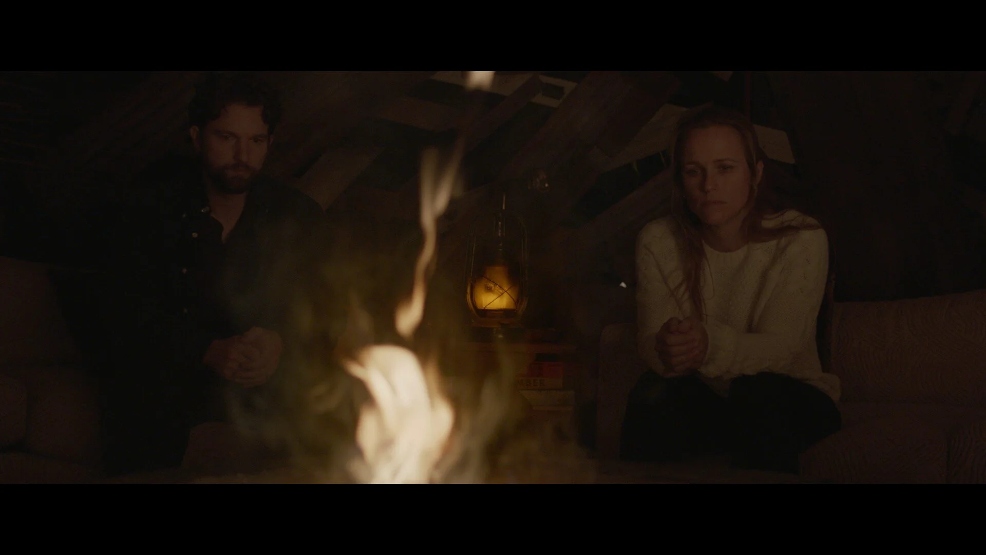Man and woman sitting in a dimly lit room around a fire, with a lantern on a table in the background.