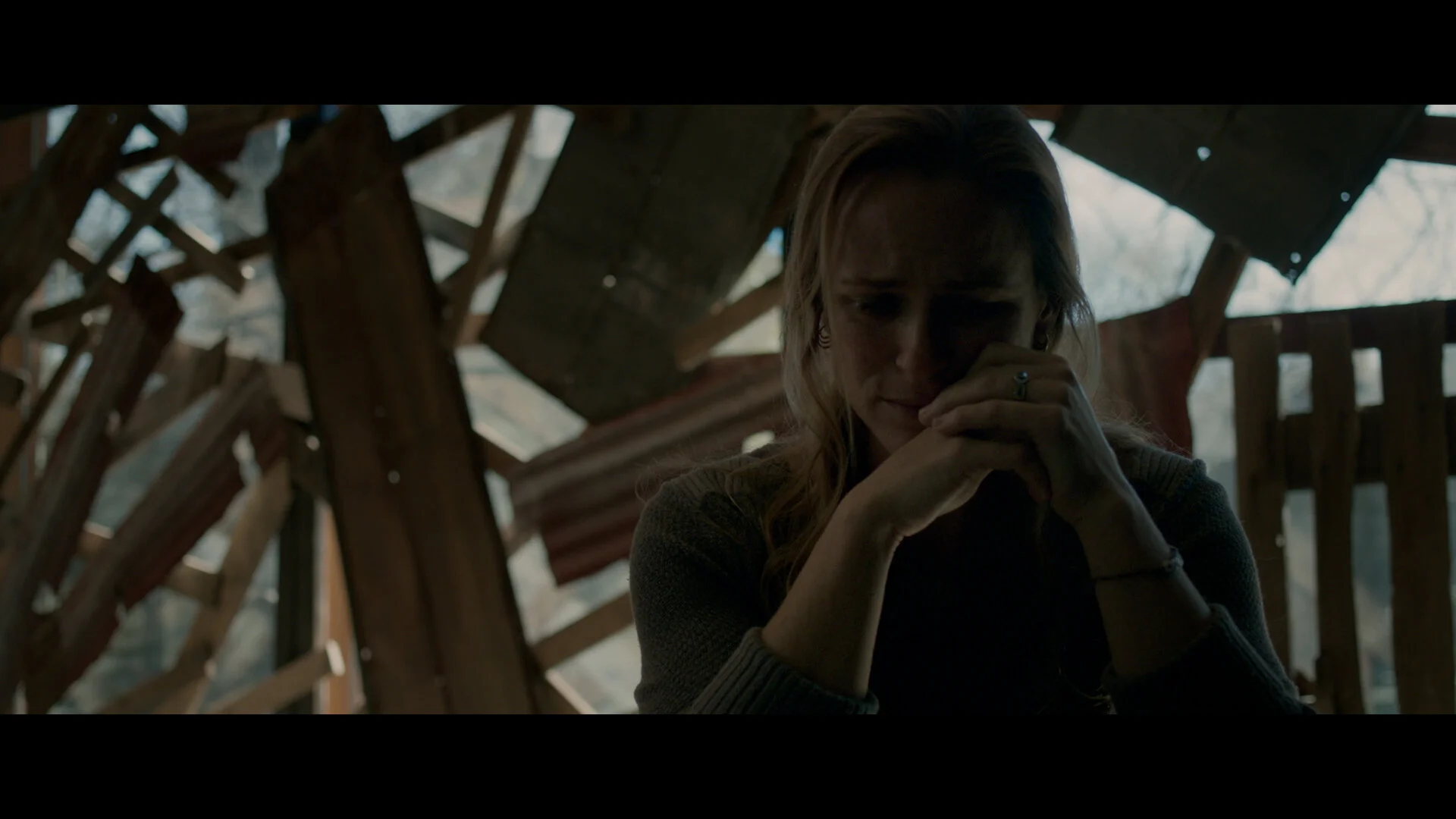 A woman sitting in a dark, wooden structure, with her hands clasped near her face, appearing to be in deep thought or distress.