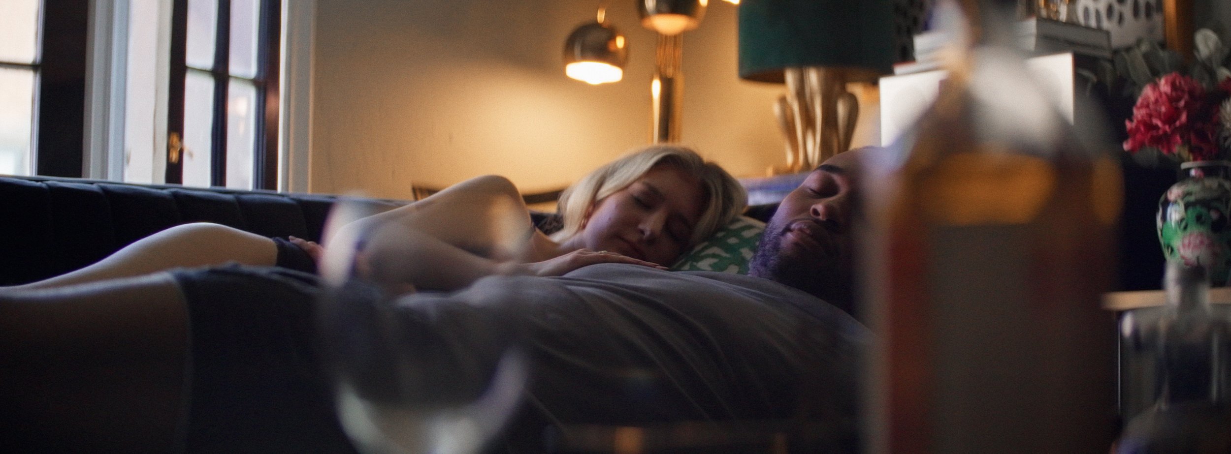 A woman and a man are sleeping together on a couch in a dimly lit room near a window, with a lamp and flowers in the background.