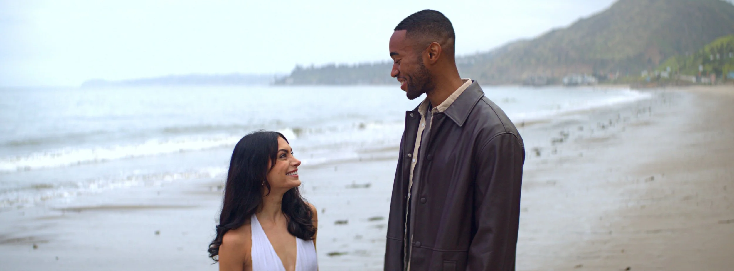 A man and woman smiling at each other on a beach, with the ocean and distant hills in the background.