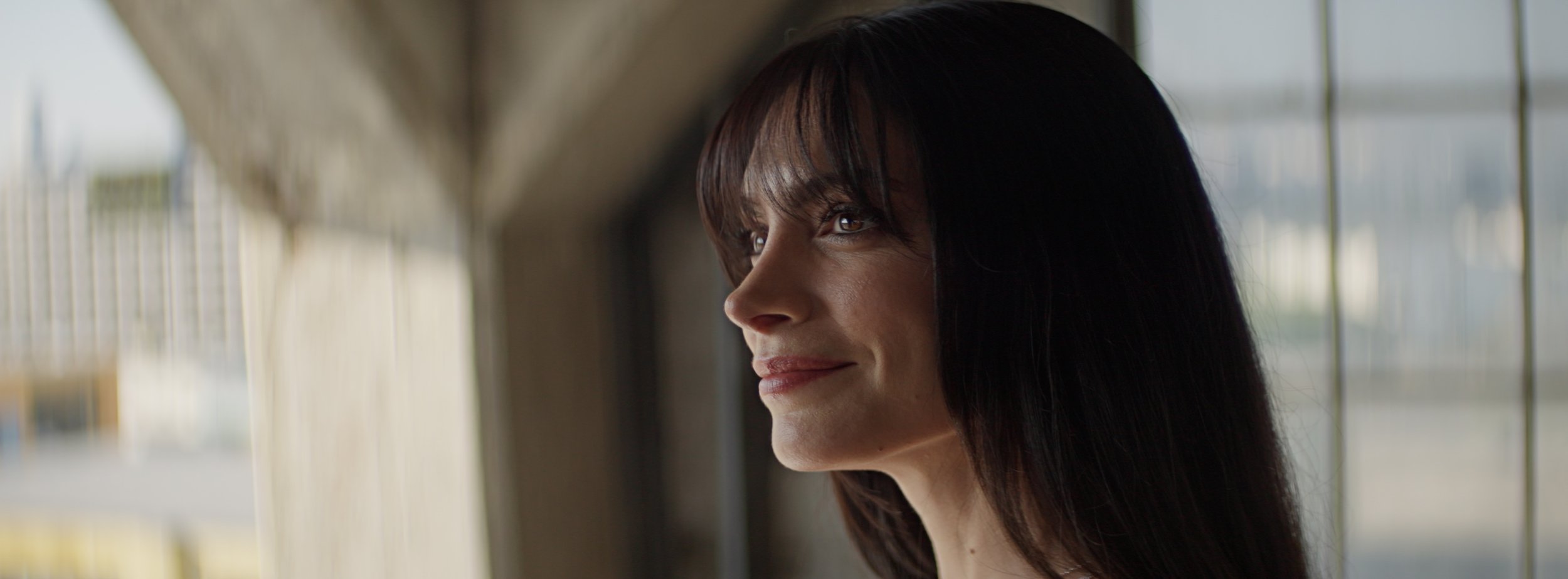 A woman with long dark hair and a slight smile looking out a window.