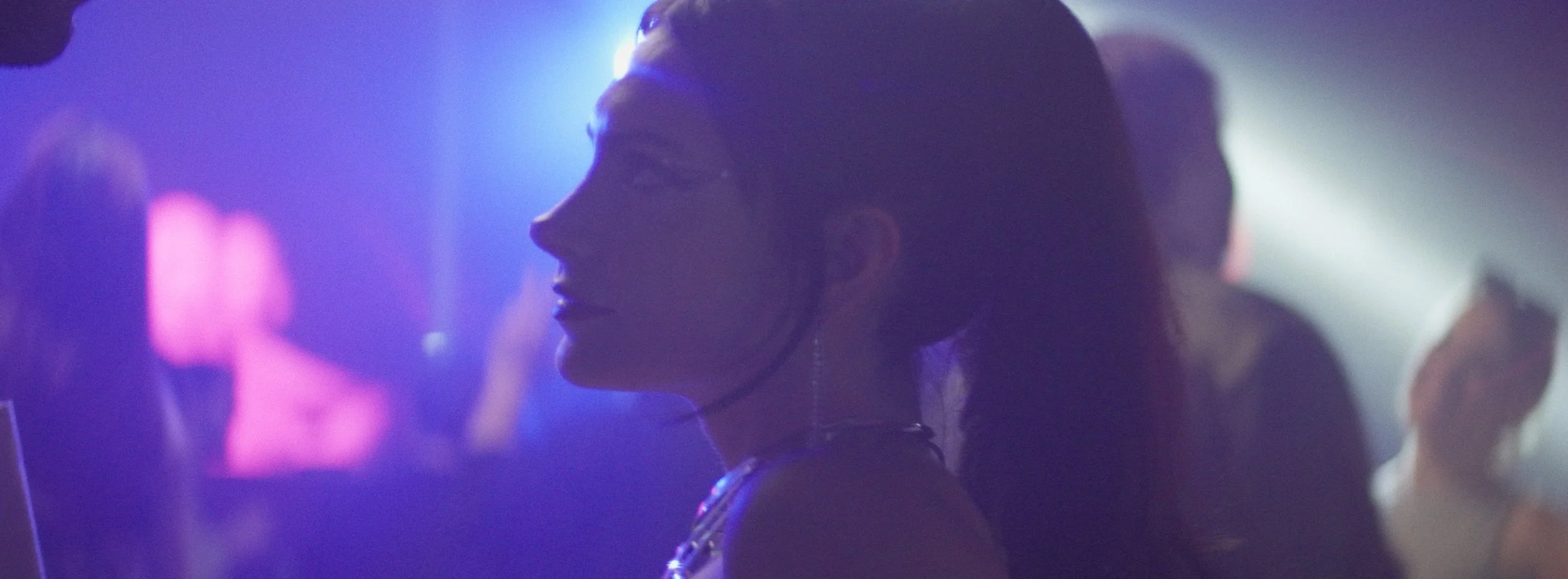 A woman with her hair in a ponytail is in a dimly lit, colorful nightclub with purple and blue lighting, surrounded by other people.