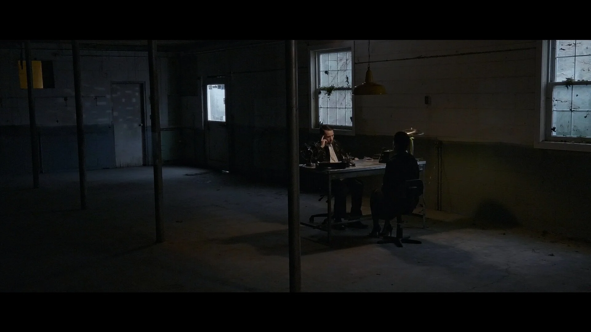 Two people sitting at a table in an old, dimly lit, industrial-style room with windows, one on the phone, the other turned away, with minimal natural light coming in.