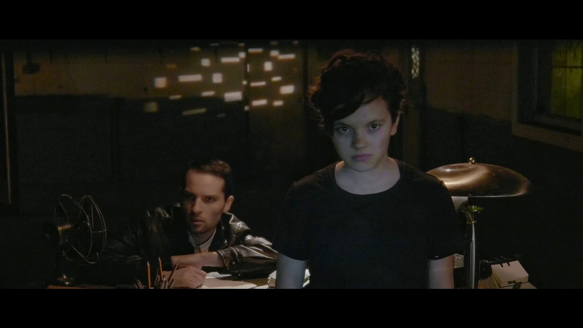A young woman with short wavy dark hair and a serious expression stands at a desk, with a man sitting behind her in a dark room with a window and blinds creating a patterned shadow in the background.