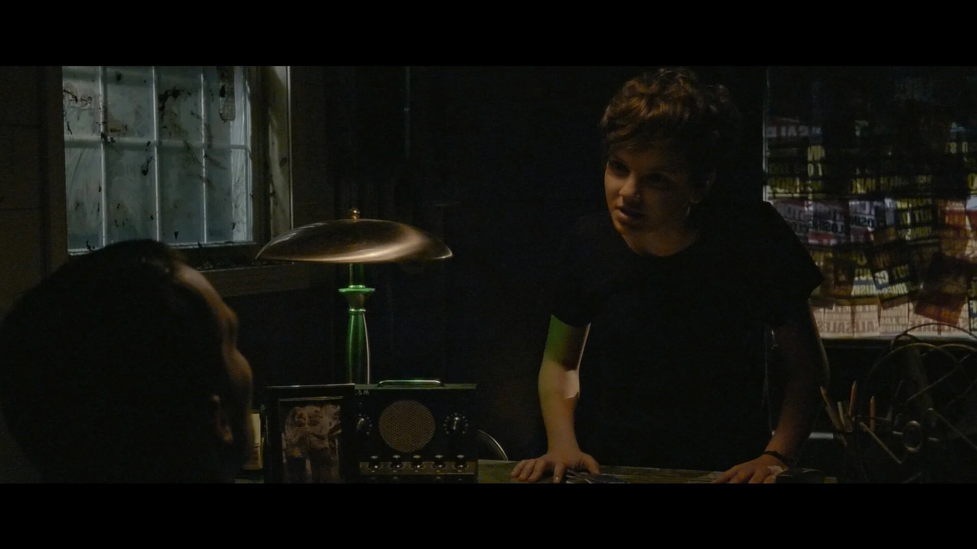 Two young men having a serious conversation inside a dimly-lit room with a window and a lamp on a desk.