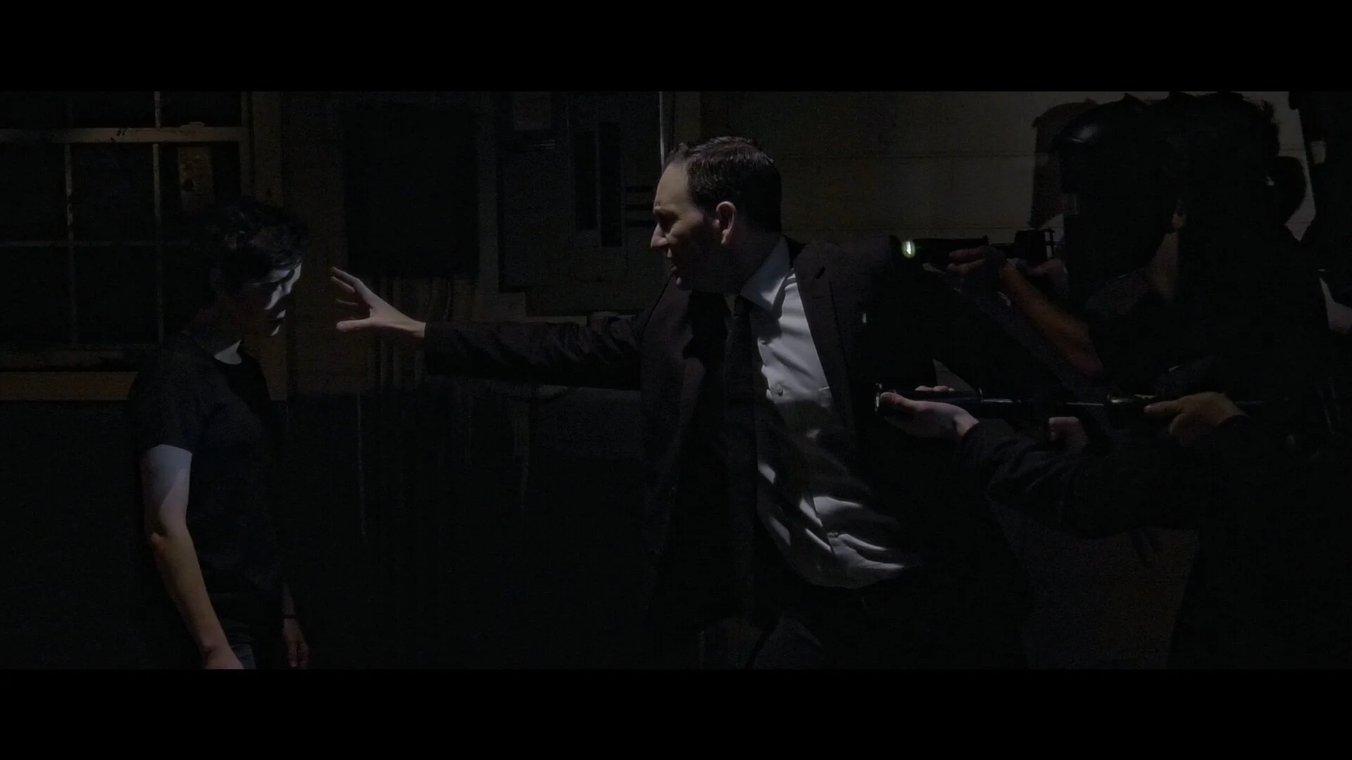 A man in a suit and tie reaches out to a young person with face paint, while surrounded by police officers aiming guns in a dark room.