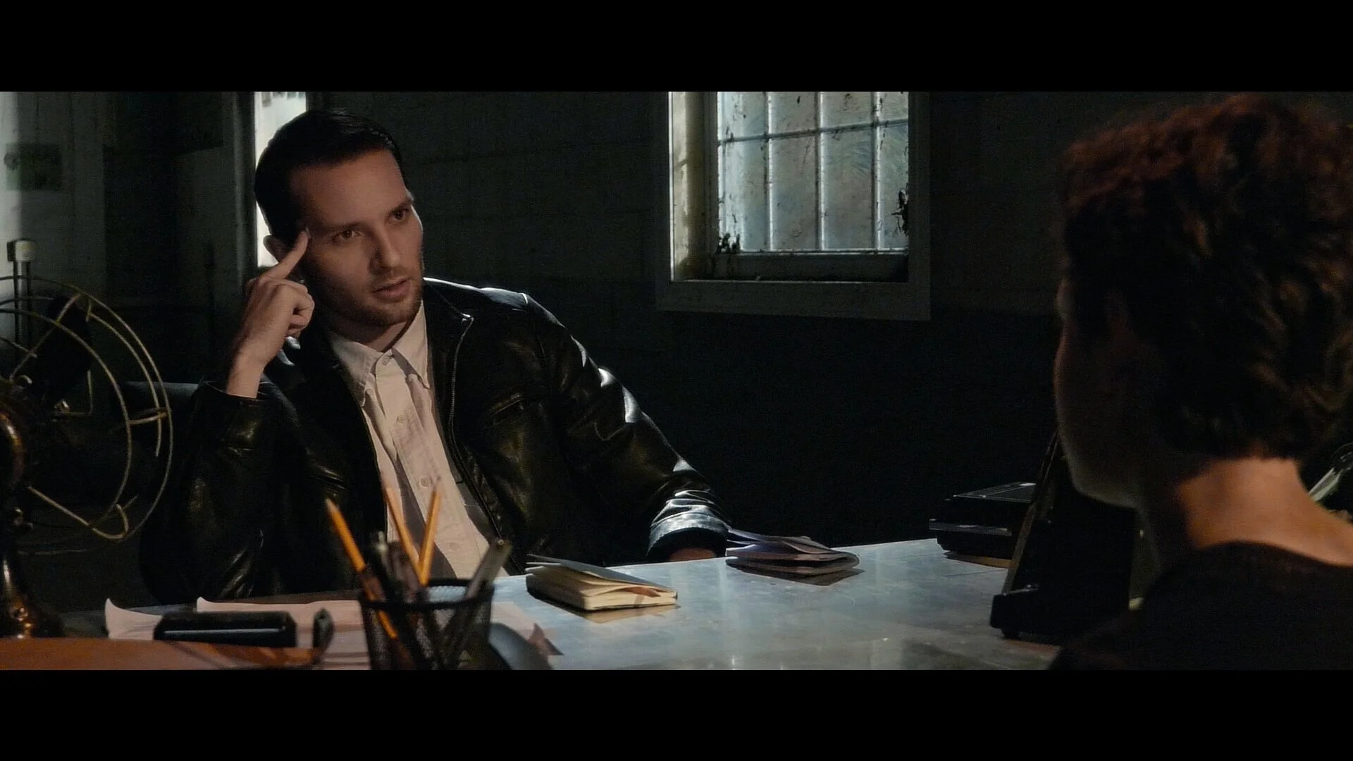 Man with dark hair and a beard sitting at a desk, talking to a woman whose back is to the camera, in a dimly lit room with a window in the background.