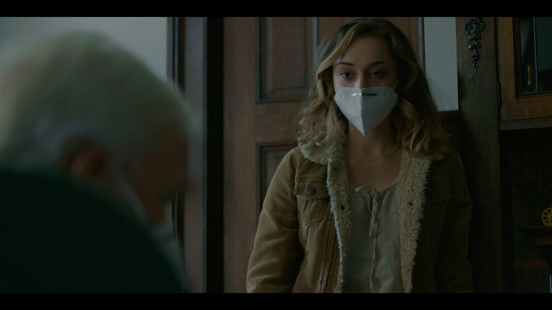 A young woman with curly hair wearing a face mask standing inside a house, talking to an elderly person with white hair, who is partially visible in the foreground.