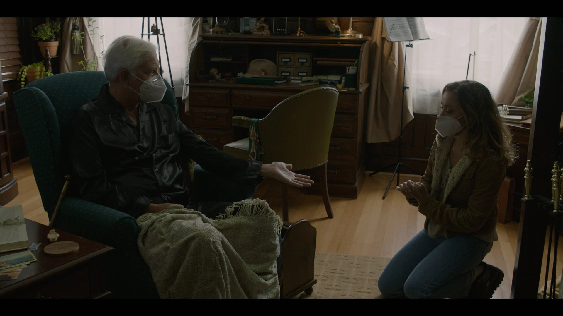 An elderly man and a young woman, both wearing masks, are engaged in a conversation inside a cozy, wood-paneled room. The man is seated in a green armchair, with his hand extended. The woman is kneeling on the floor facing him, with her hands clasped