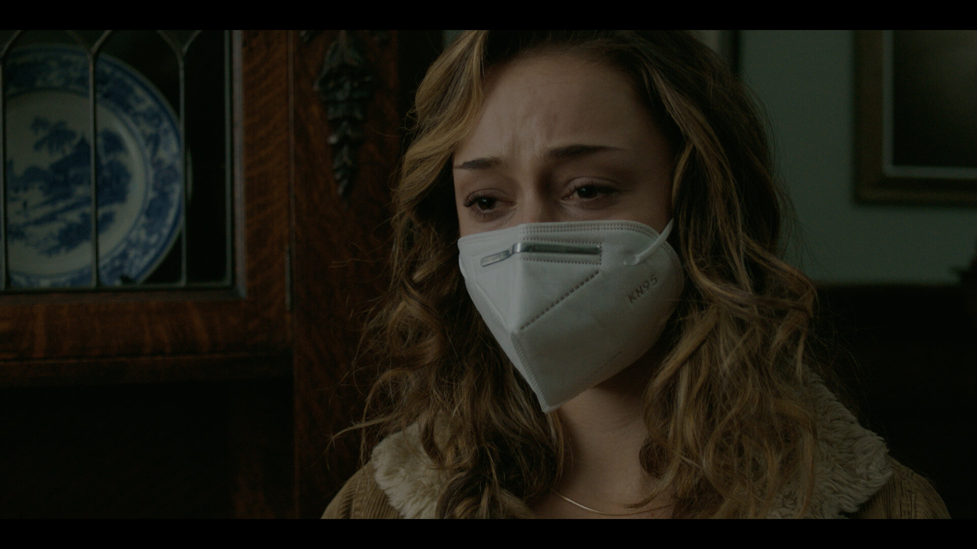 A woman with curly hair wearing a KN95 face mask in a room with wooden furniture and framed artwork on the wall.