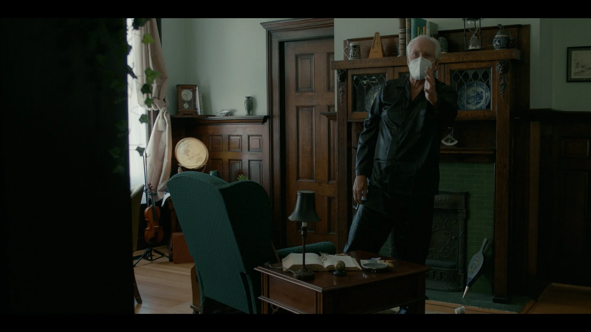An elderly man wearing a face mask standing in a living room with wooden furniture and a small table with an open book, a lamp, and a remote control, next to a green armchair and a fireplace with decorative items on the mantel.