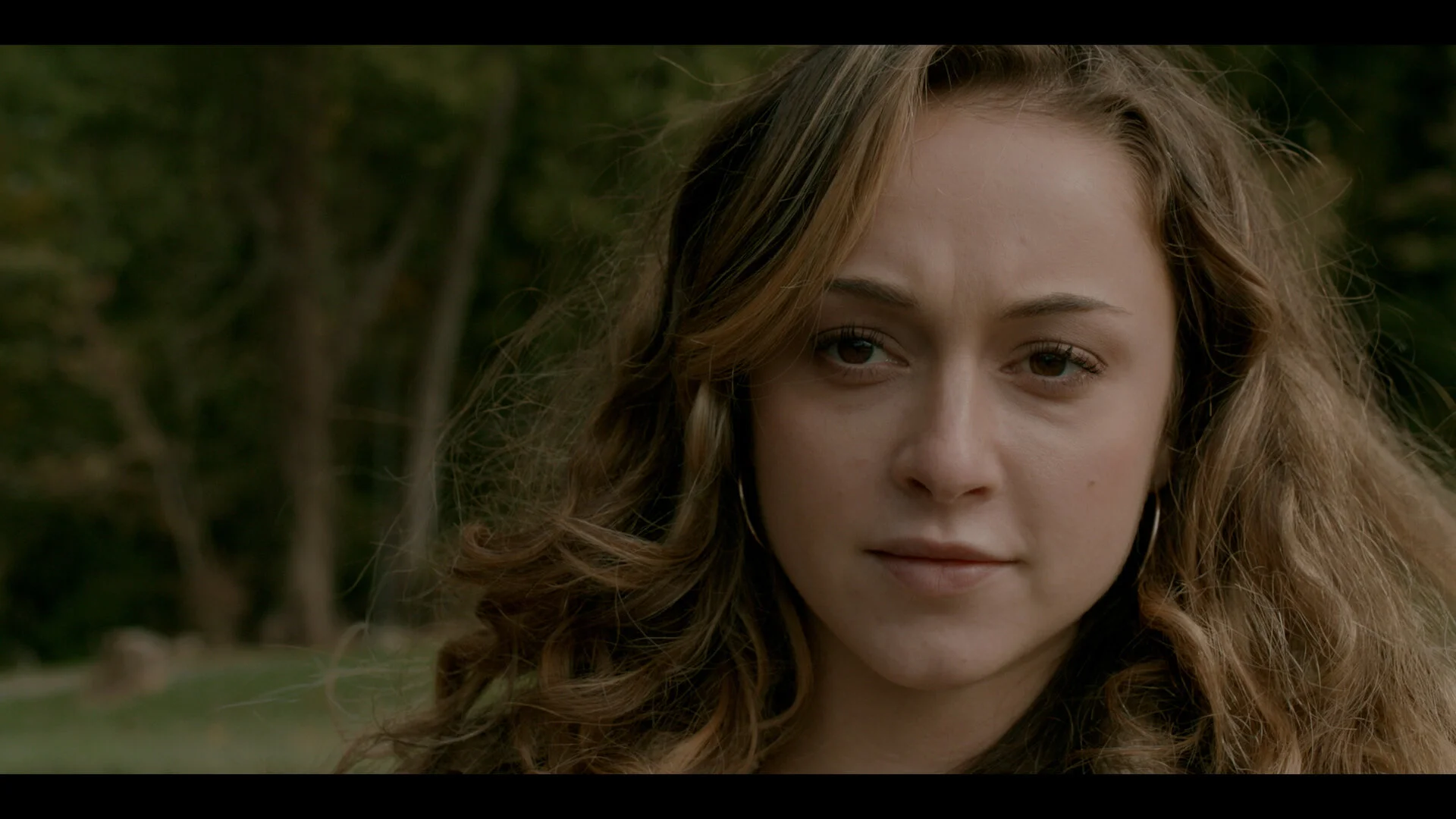 Close-up of a young woman with curly light brown hair and hoop earrings outdoors, with trees and greenery in the background.