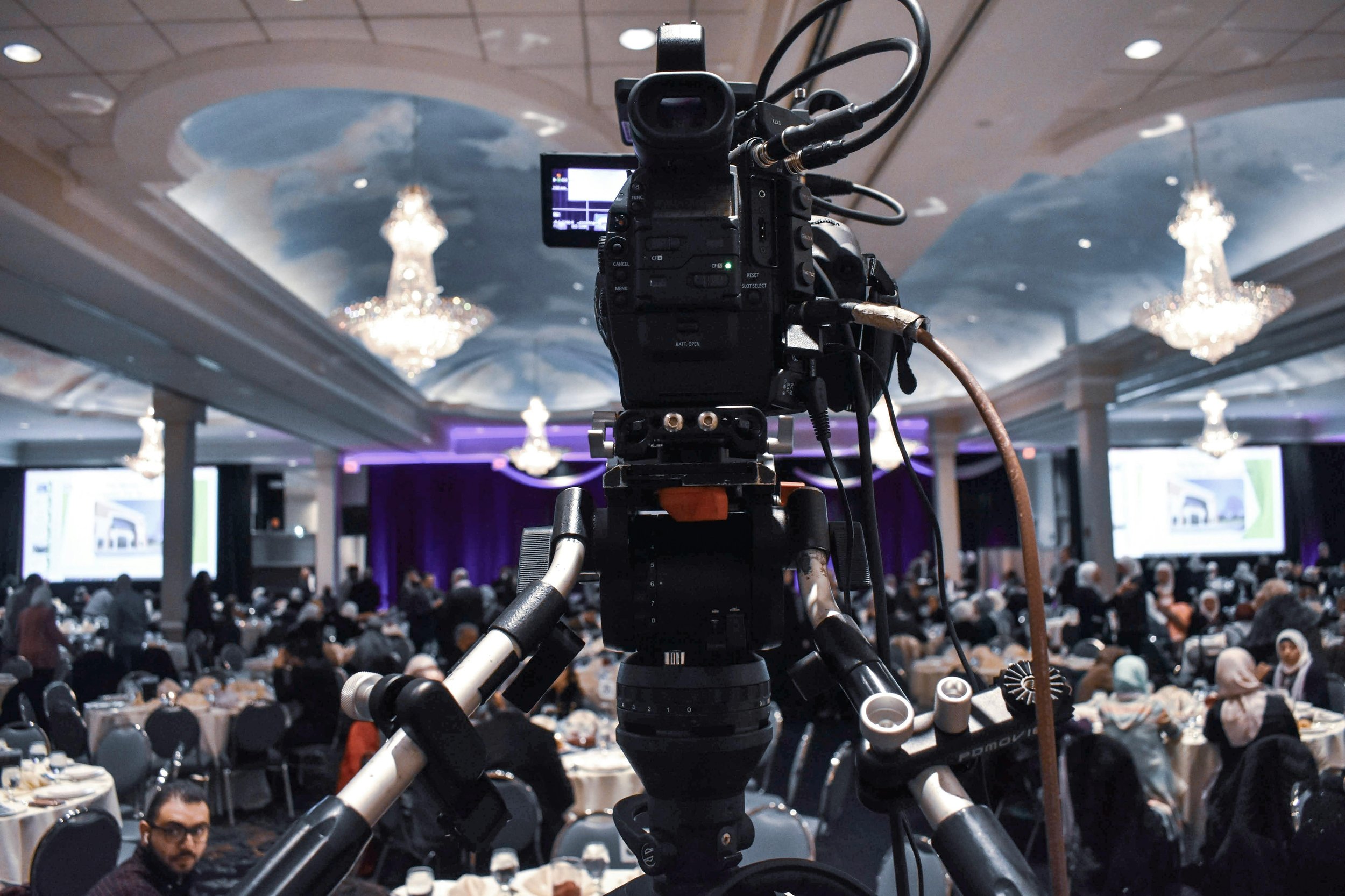 Camera setup pointed at an audience in a large banquet hall with chandeliers, where people are seated at round tables, some of whom are looking toward the stage or camera.
