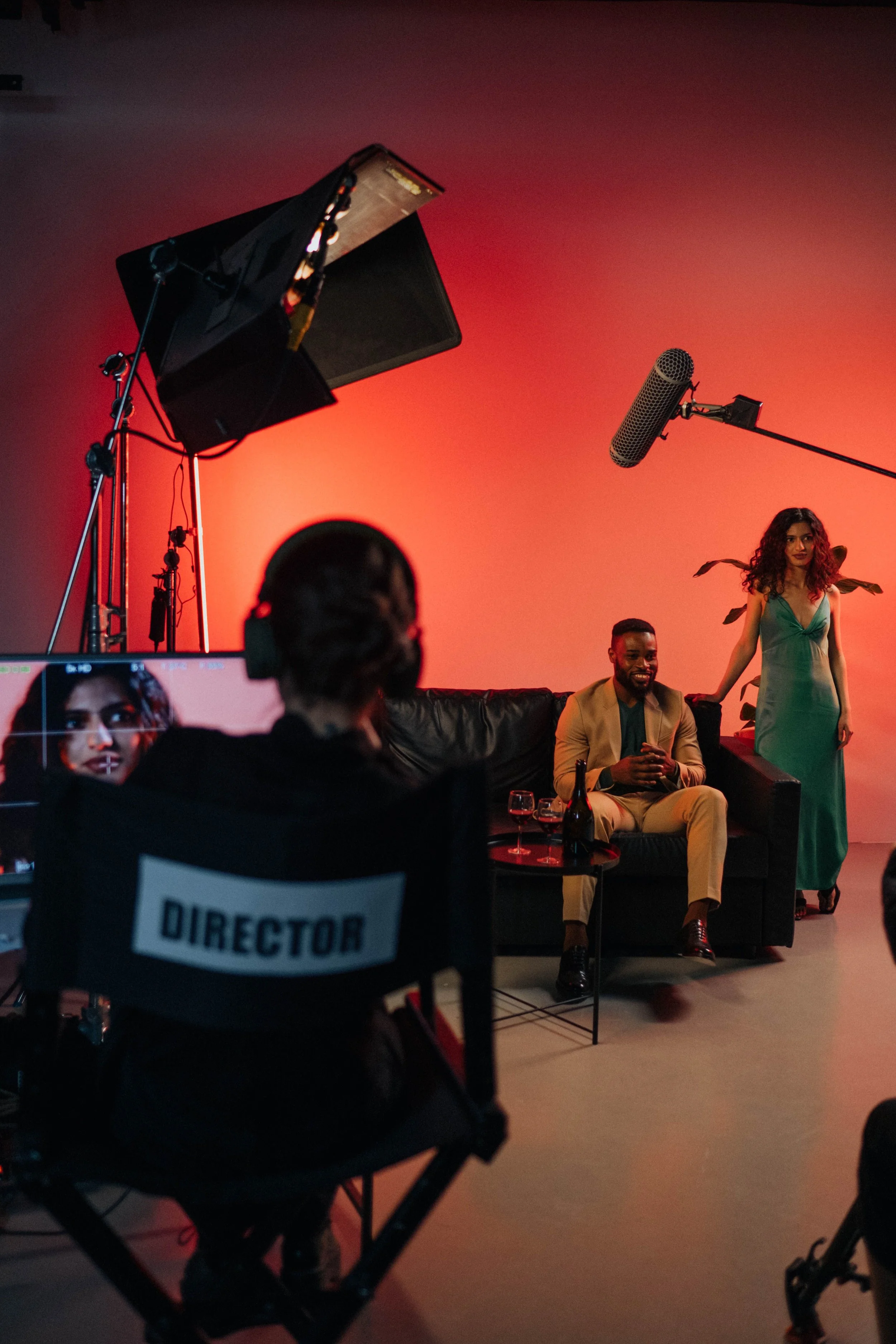 A behind-the-scenes view of a film or music video shoot with lighting equipment, a red background, and three people in the scene. One person is sitting on a black couch, another is standing, and a third person's back is visible with a 'Director' chair.