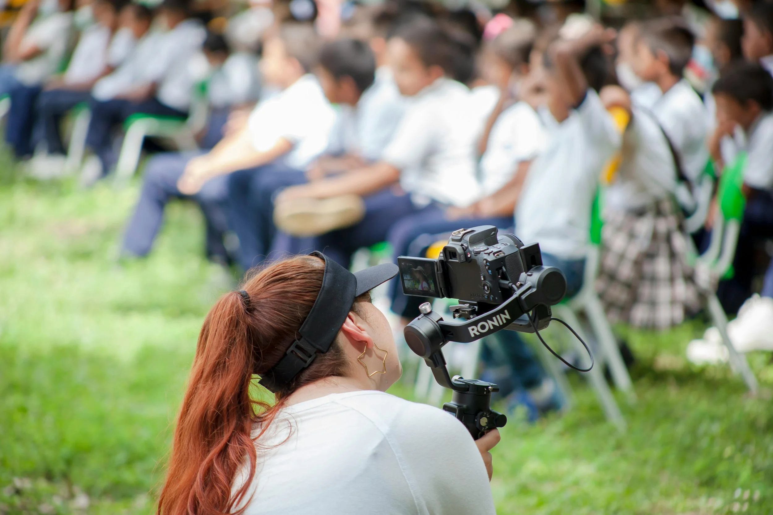A woman with long red hair is operating a professional video camera outdoors during the daytime. In the background, a group of children are seated on chairs in a row, wearing white shirts and dark pants or skirts, with some wearing glasses or face masks, attending an outdoor event or gathering on a grassy area.