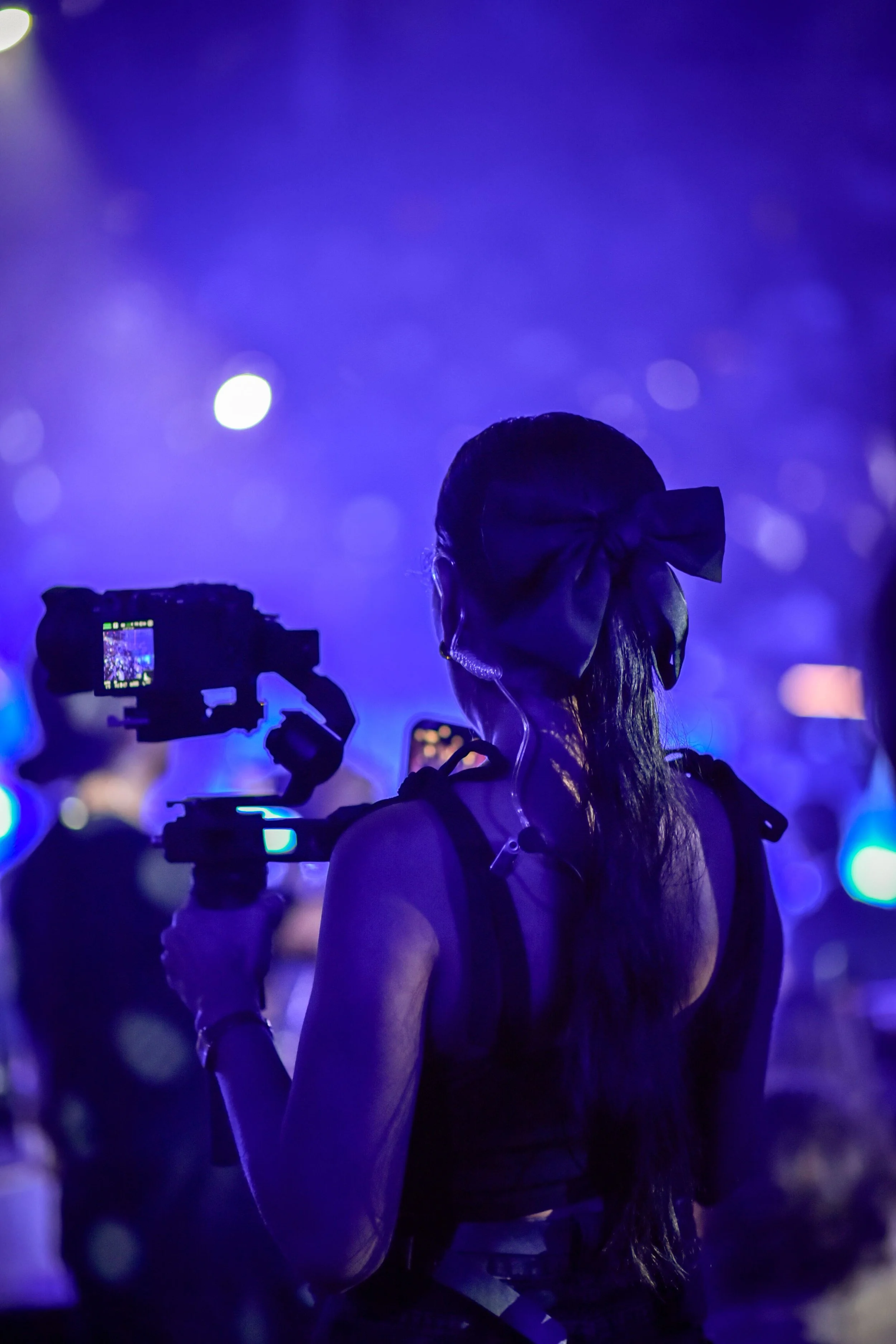 A woman with long hair tied with a large bow, standing in a dark, vibrant purple-lit environment, possibly at a concert or event, with people and cameras capturing the scene.