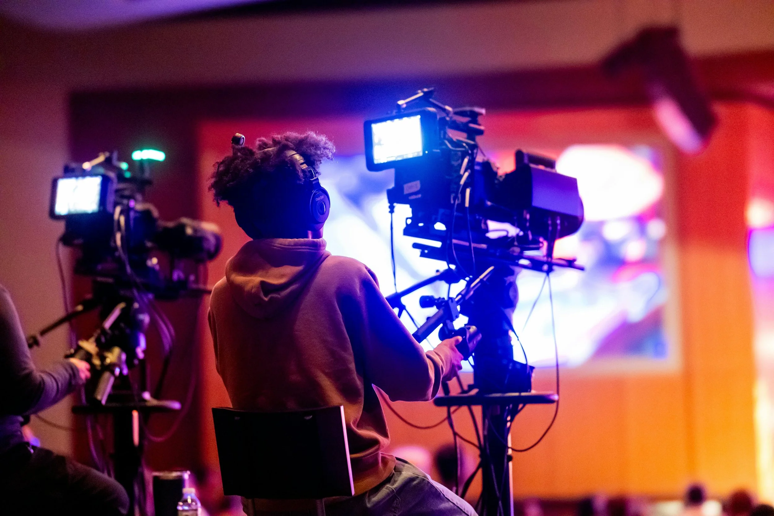 A person operating professional video camera equipment in a dimly lit environment with colorful lights.