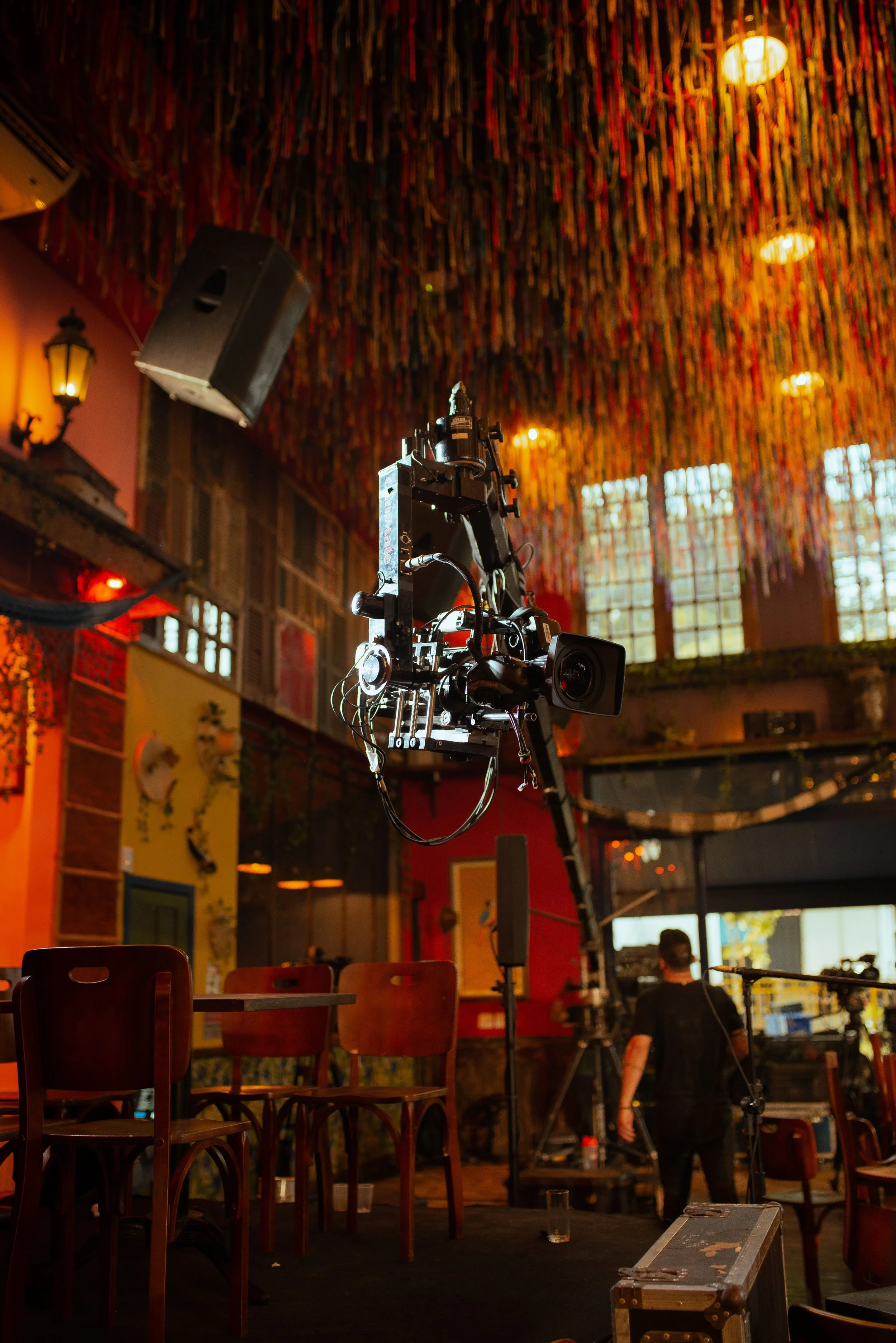 Inside a venue with a stage setup, a camera on a tripod, chairs in the foreground, warm lighting, colorful decorations hanging from the ceiling, and a person working near the stage.