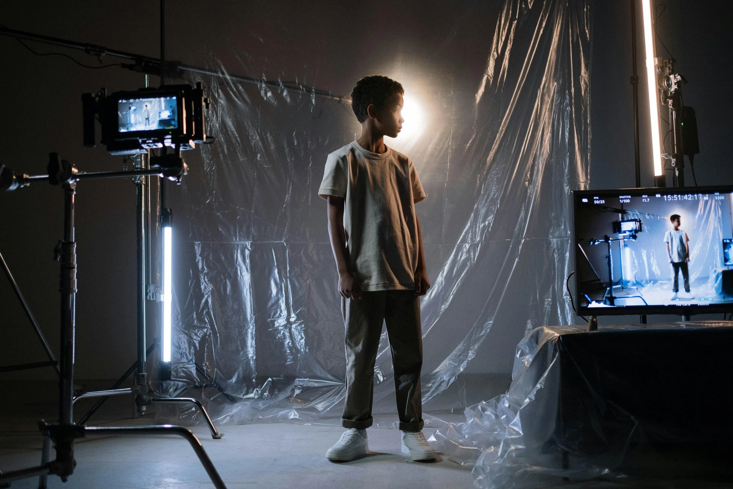A person standing in a photo studio with film equipment, a monitor, and lighting, with a backlit backdrop and plastic-covered floor.