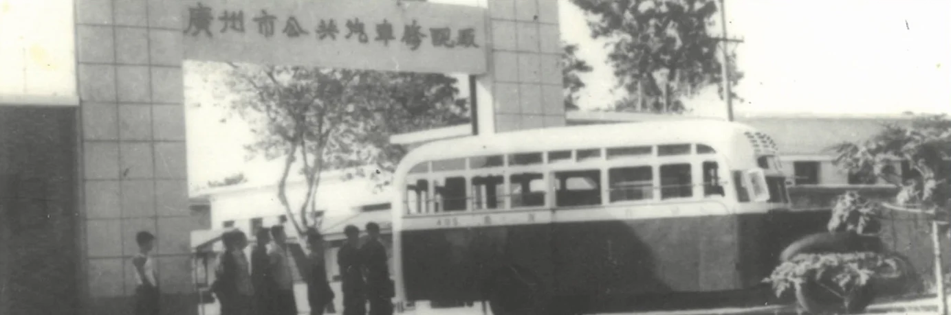 Black and white photo of a vintage bus parked in front of a building with Chinese characters on the sign, and a few people standing nearby.