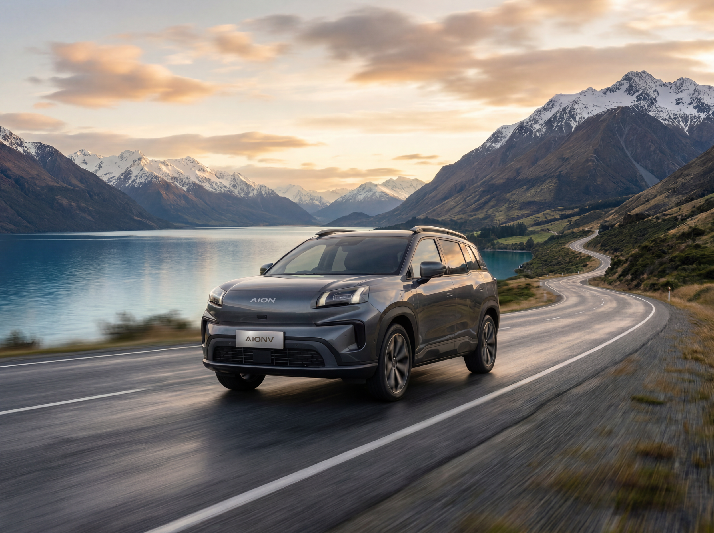 A gray electric car driving on a winding mountain road beside a lake, with snow-capped mountains and a partly cloudy sky in the background.