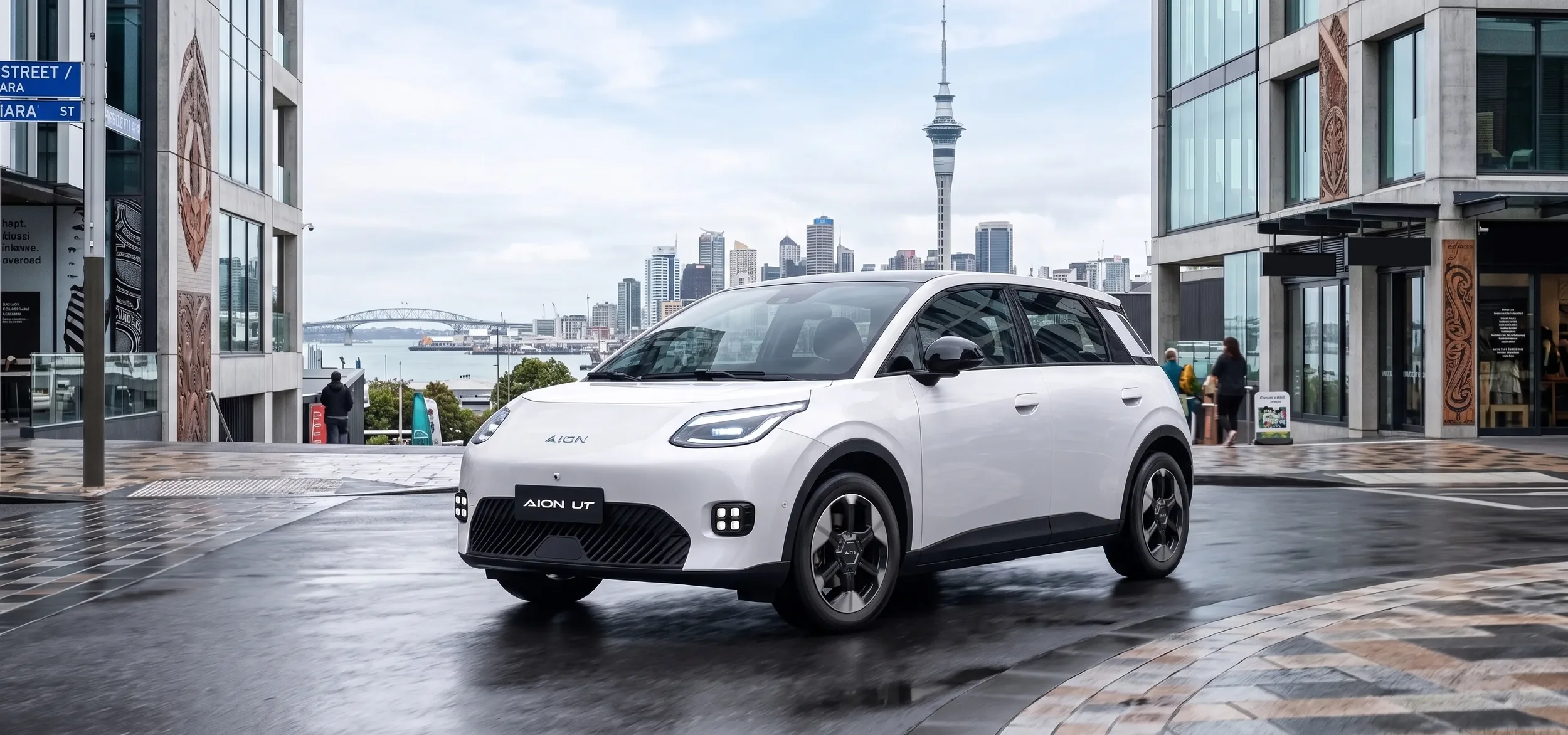 A white compact electric car on a city street with modern buildings and a view of a skyline and bridge in the background.