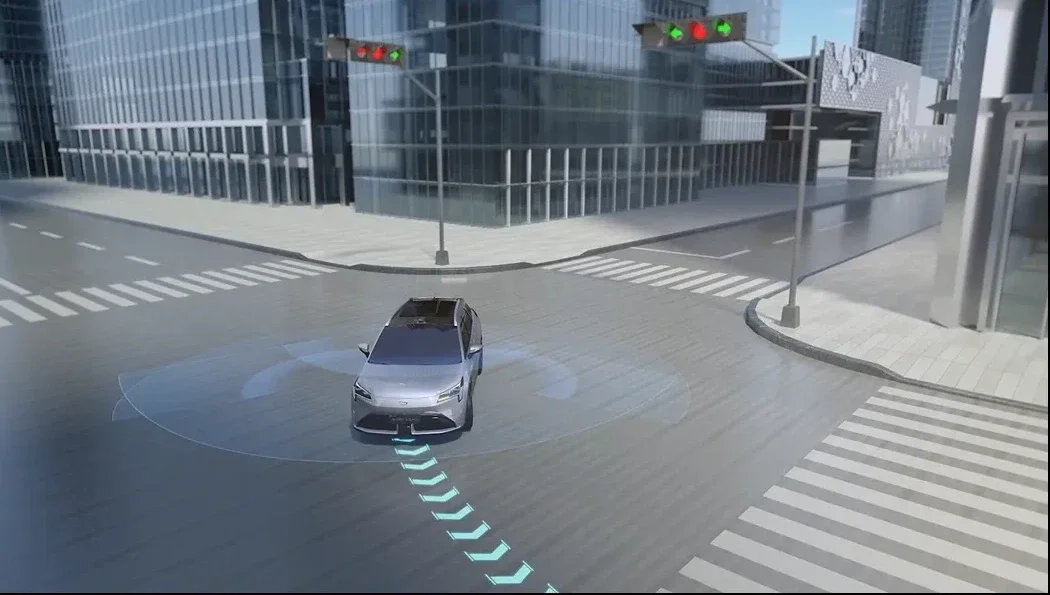 A silver autonomous vehicle stopped at an intersection with green traffic lights in an urban setting.