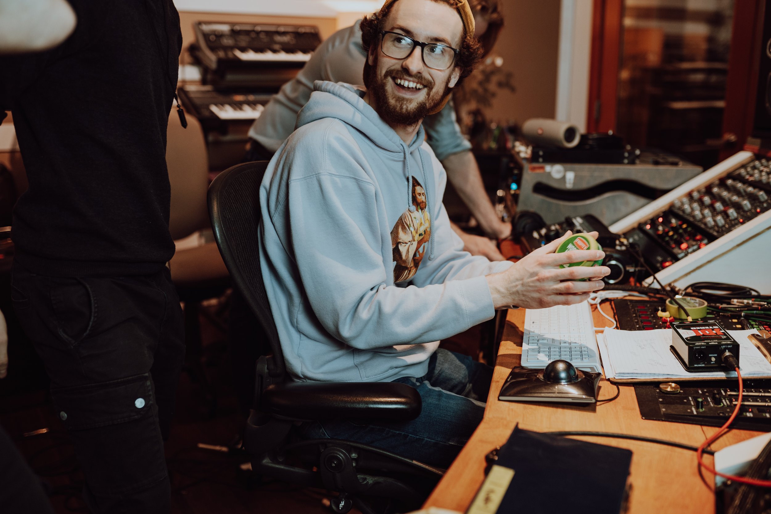 A man with glasses, a beard, and a hoodie with a Jesus print, smiling and holding a small green object, sitting at a workstation with electronic equipment and musical instruments in a recording studio. There are other people and equipment in the background.