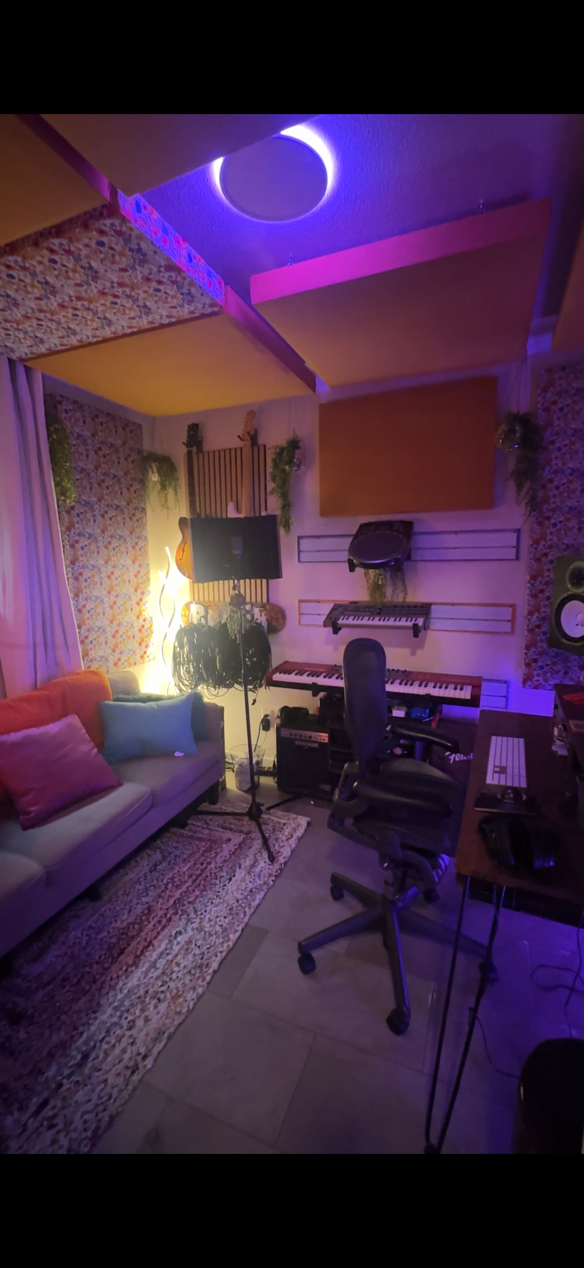 A music recording studio with a colorful acoustic panel ceiling, a couch with pillows, a keyboard, studio monitors, microphones, and various music equipment illuminated by purple and yellow lighting.
