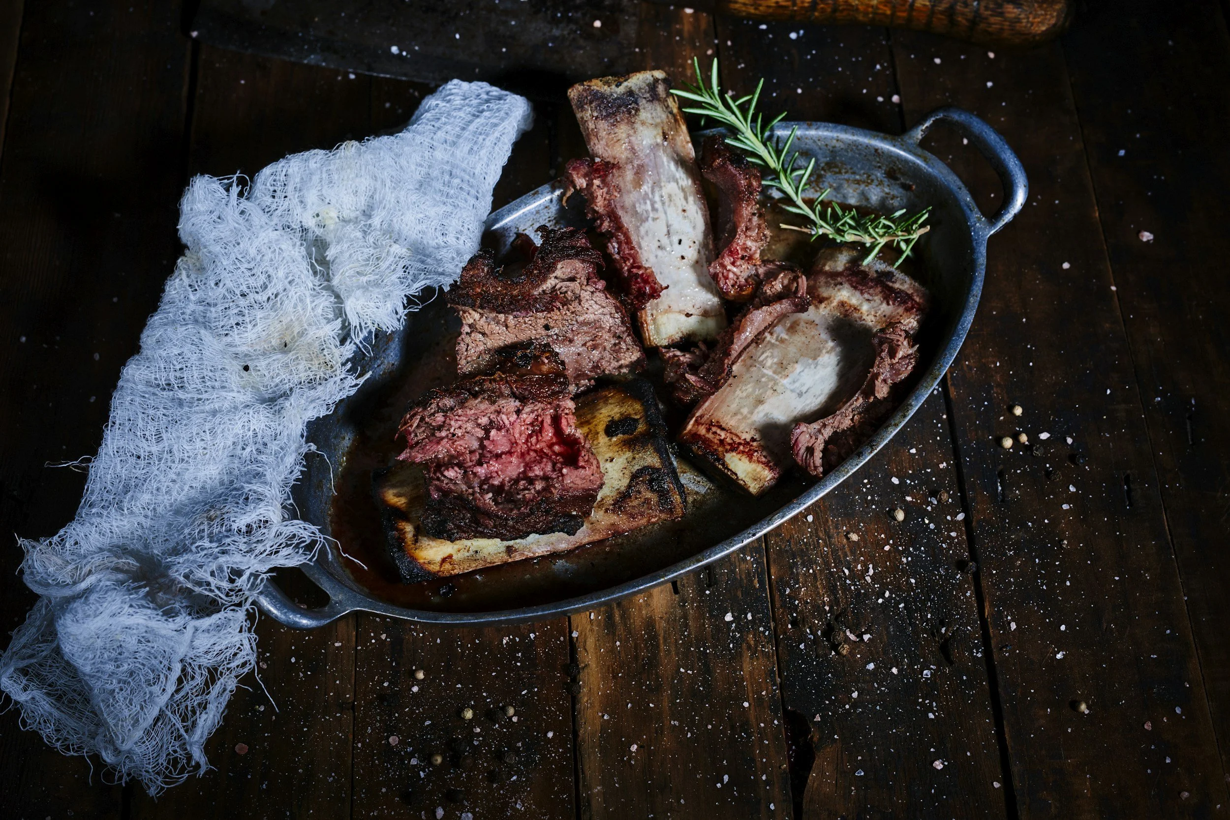 Cooked beef short ribs with sprig of rosemary on a metal platter, a white cheesecloth, and scattered seasoning on a dark wooden surface.