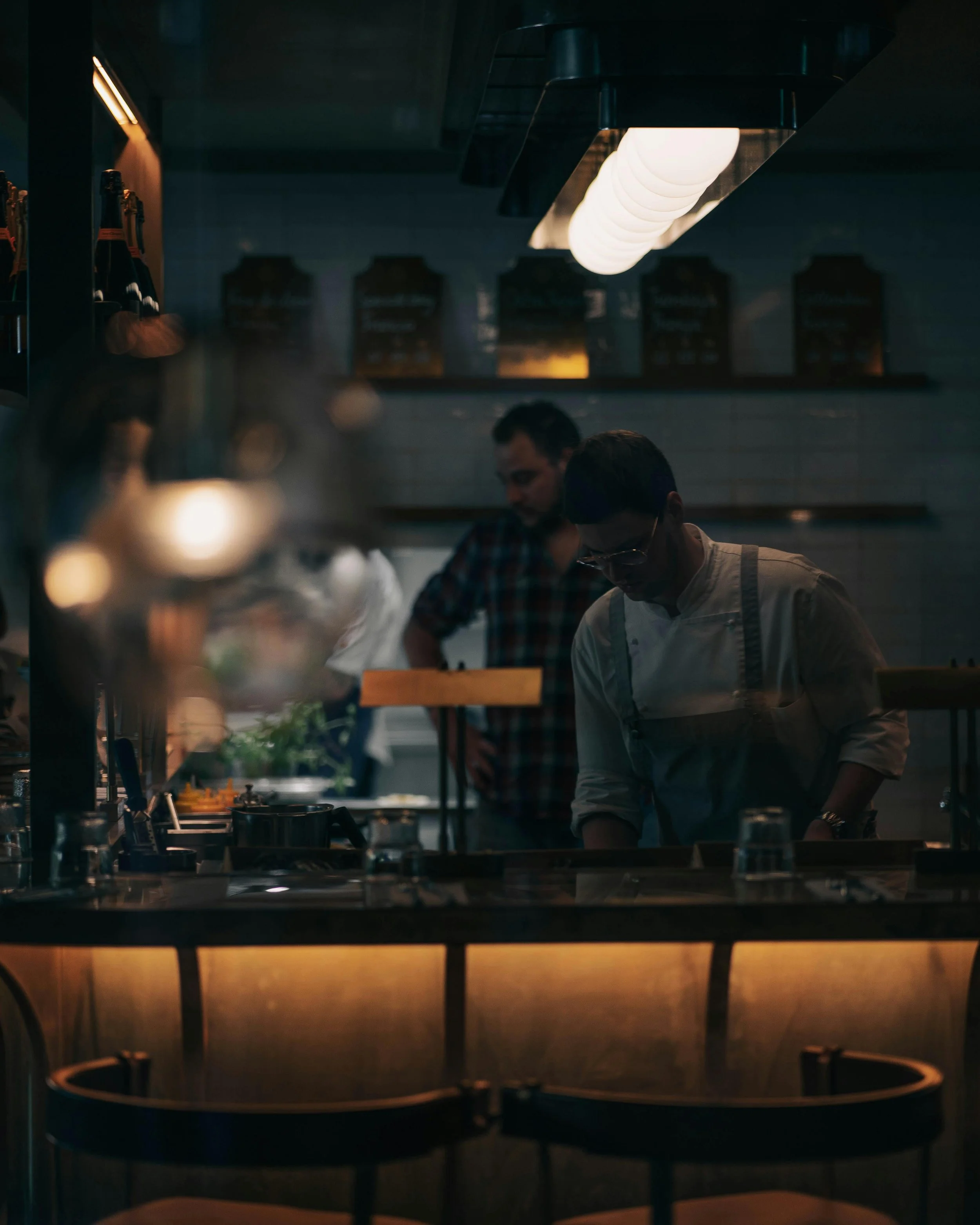 A dimly lit kitchen with two men preparing food. Overhead, a row of white lights are hanging. The background shows a menu on the wall and bottles on shelves. The foreground features a glass of water and a blurred reflection of the scene.