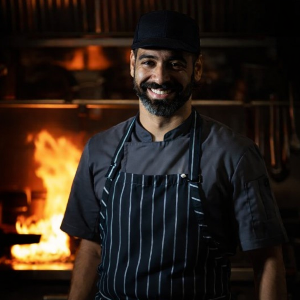 A smiling male chef in a black hat and striped apron standing in a restaurant kitchen with a fire in the background. Executive Chef Federico Pitasi, F&B and kitchen operations consultant at HPK Consultancy, in a commercial kitchen in Australia.