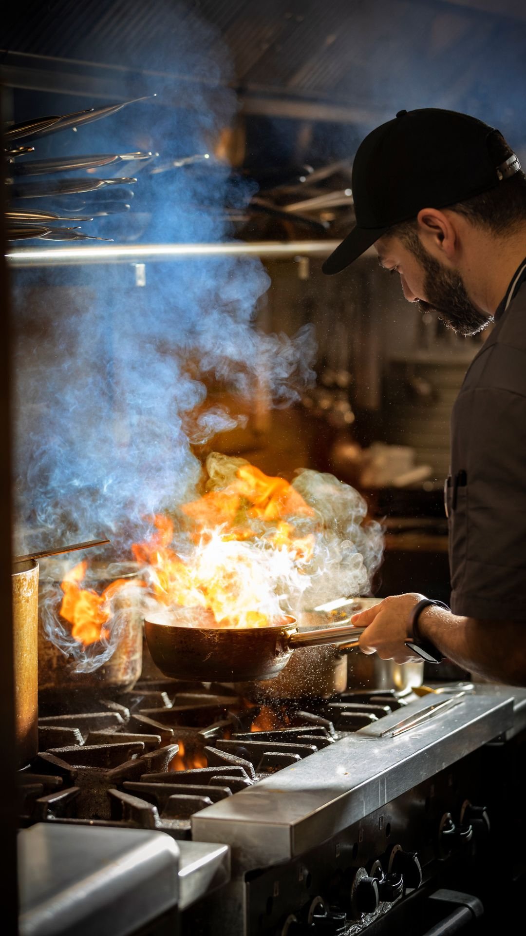 A chef wearing a black cap and gray shirt cooking on a stovetop with flames and smoke coming from a pan.
