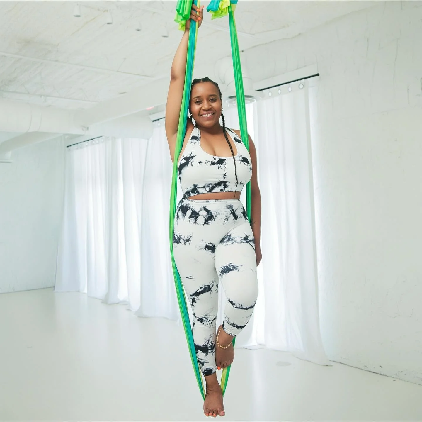 A smiling woman in a black and white patterned workout outfit, holding aerial silk fabric in green and blue, standing in a bright, white room with curtains.