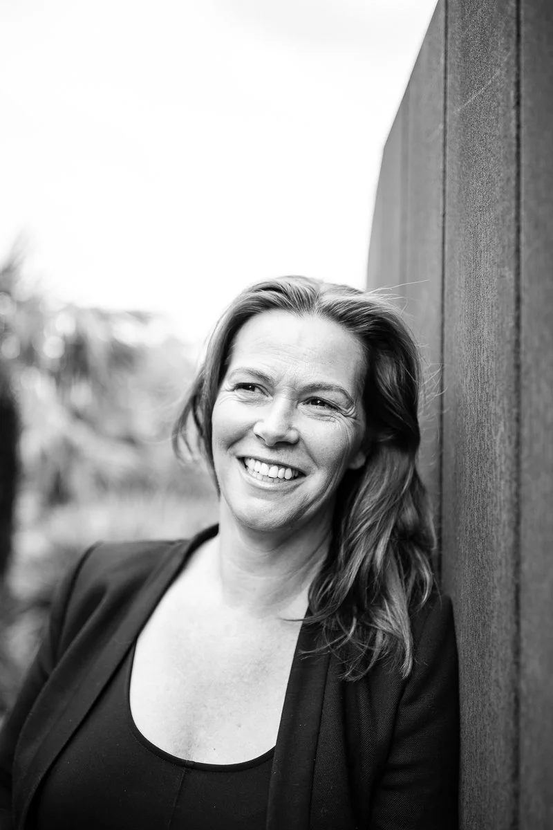 Black and white photo of a woman smiling, leaning against a wooden wall outdoors.