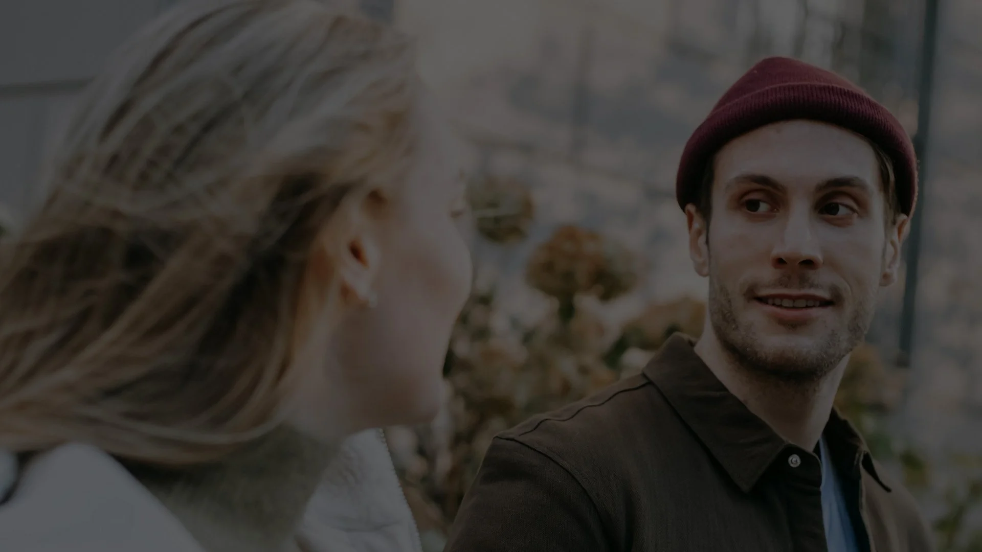A young man with a maroon beanie and dark jacket speaking with a woman with long, light brown hair, in an outdoor setting with autumn foliage.
