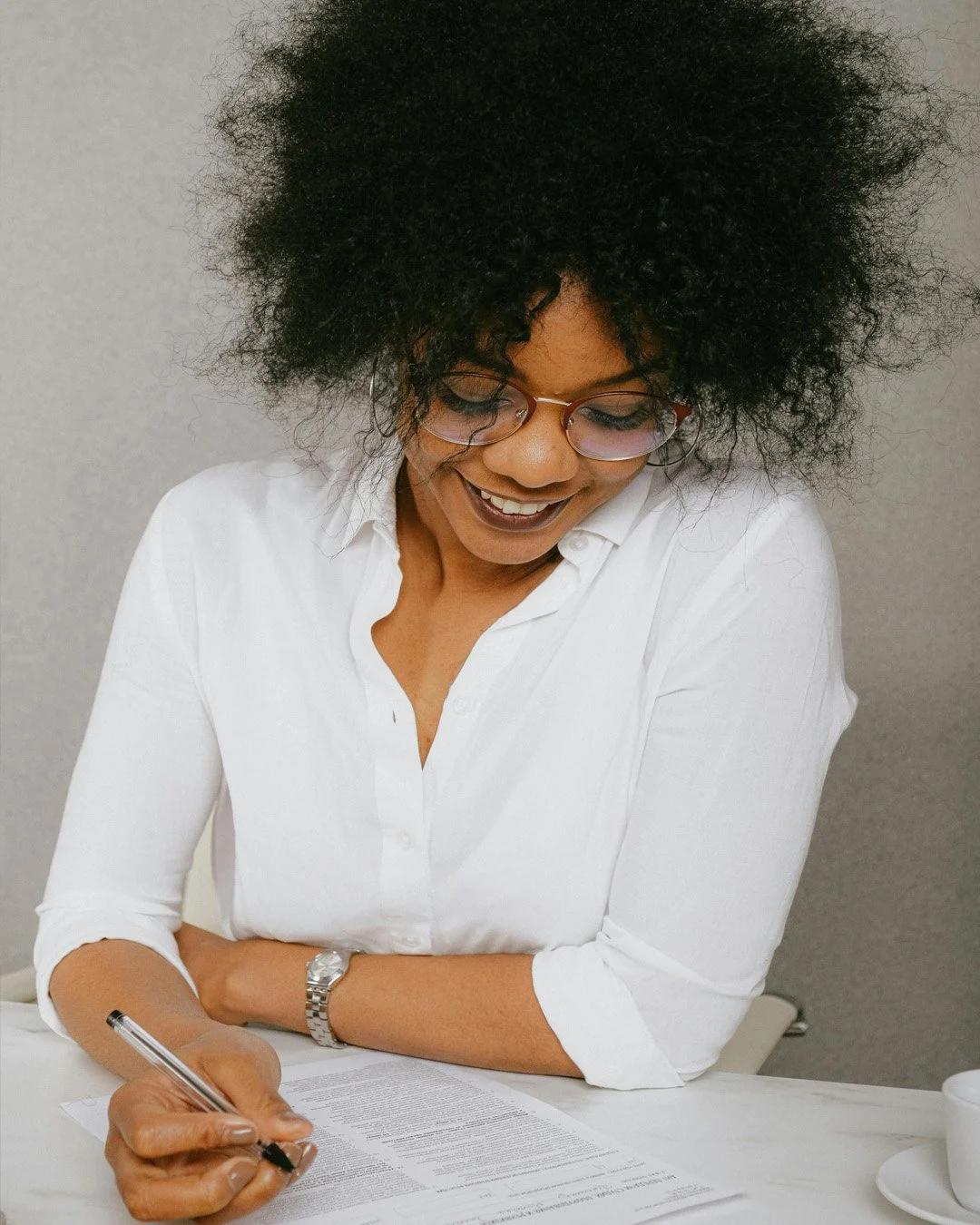 A woman with curly black hair, glasses, and a white shirt, smiling while writing on a document at a desk.